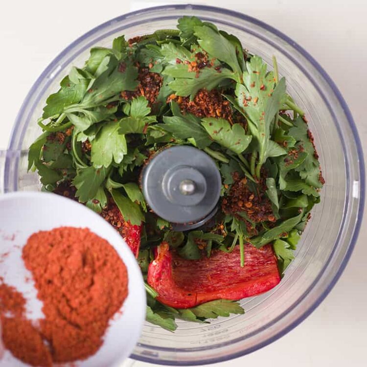 Fresh parsley, red pepper flakes, and a red bell pepper in a food processor for chopped herb and spice mixture.