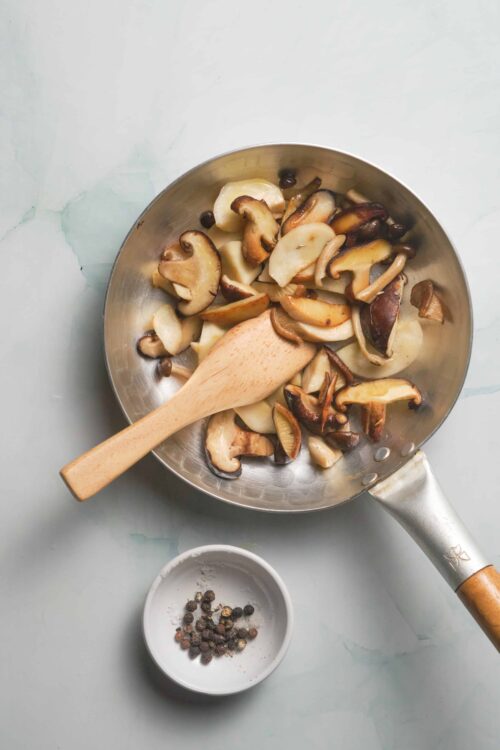 Sautéed mixed mushrooms in a stainless steel skillet with peppercorns, ready for cooking or serving.