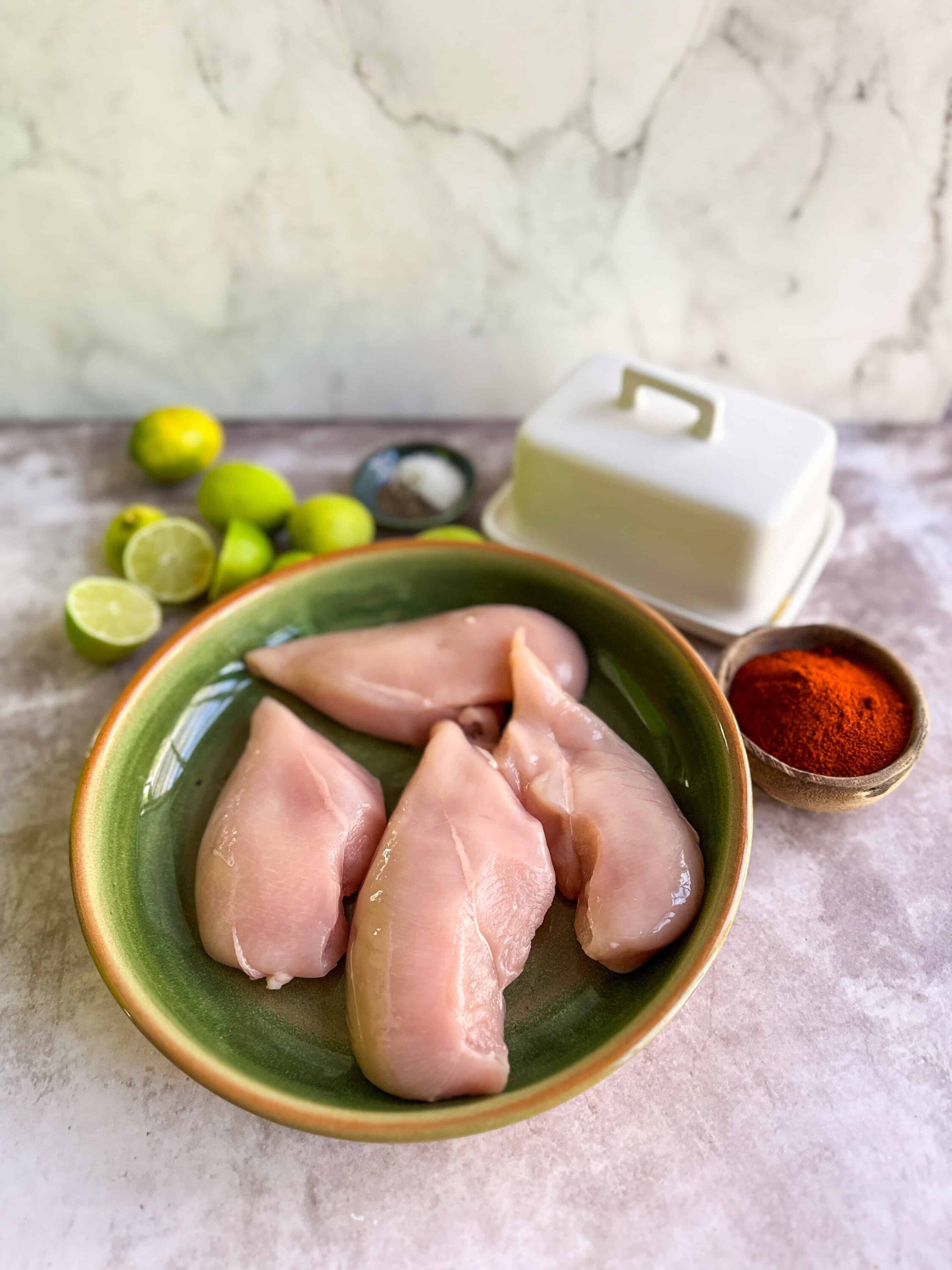 Raw chicken breasts in a green bowl with limes, spices, and butter, ready for baking or cooking.