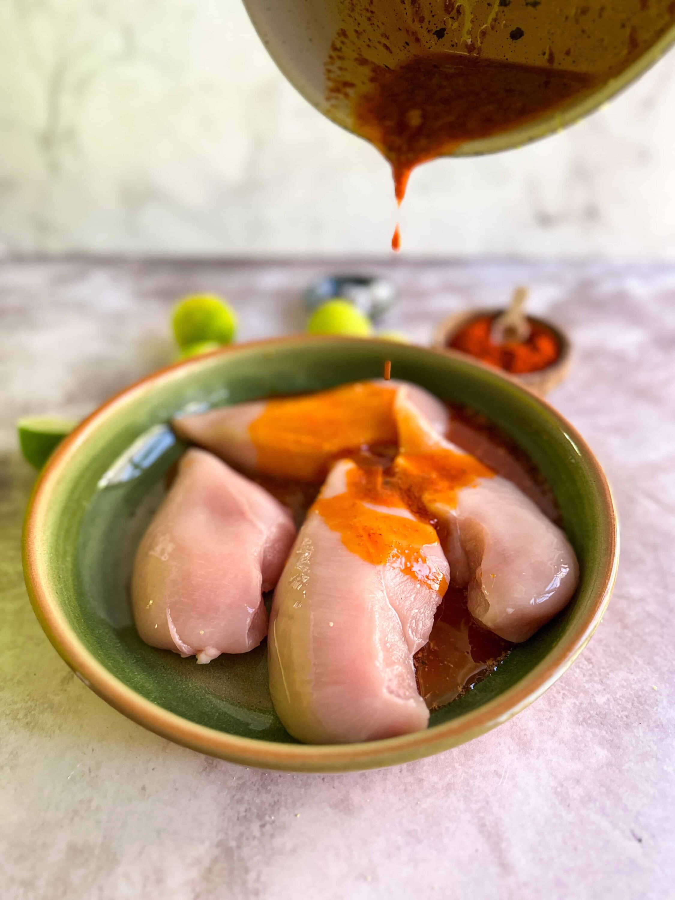 Fresh raw chicken breasts in a green bowl with marinade being poured over.