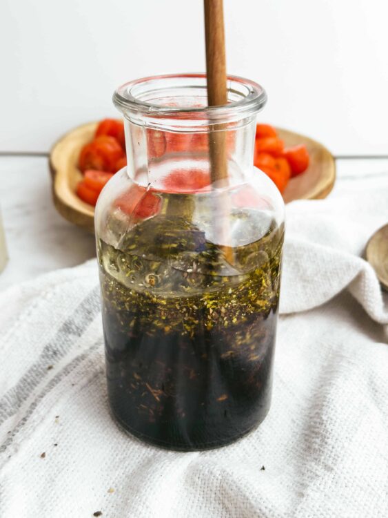Baked Bree homemade herb-infused olive oil in a glass jar with fresh chopped tomatoes in the background.