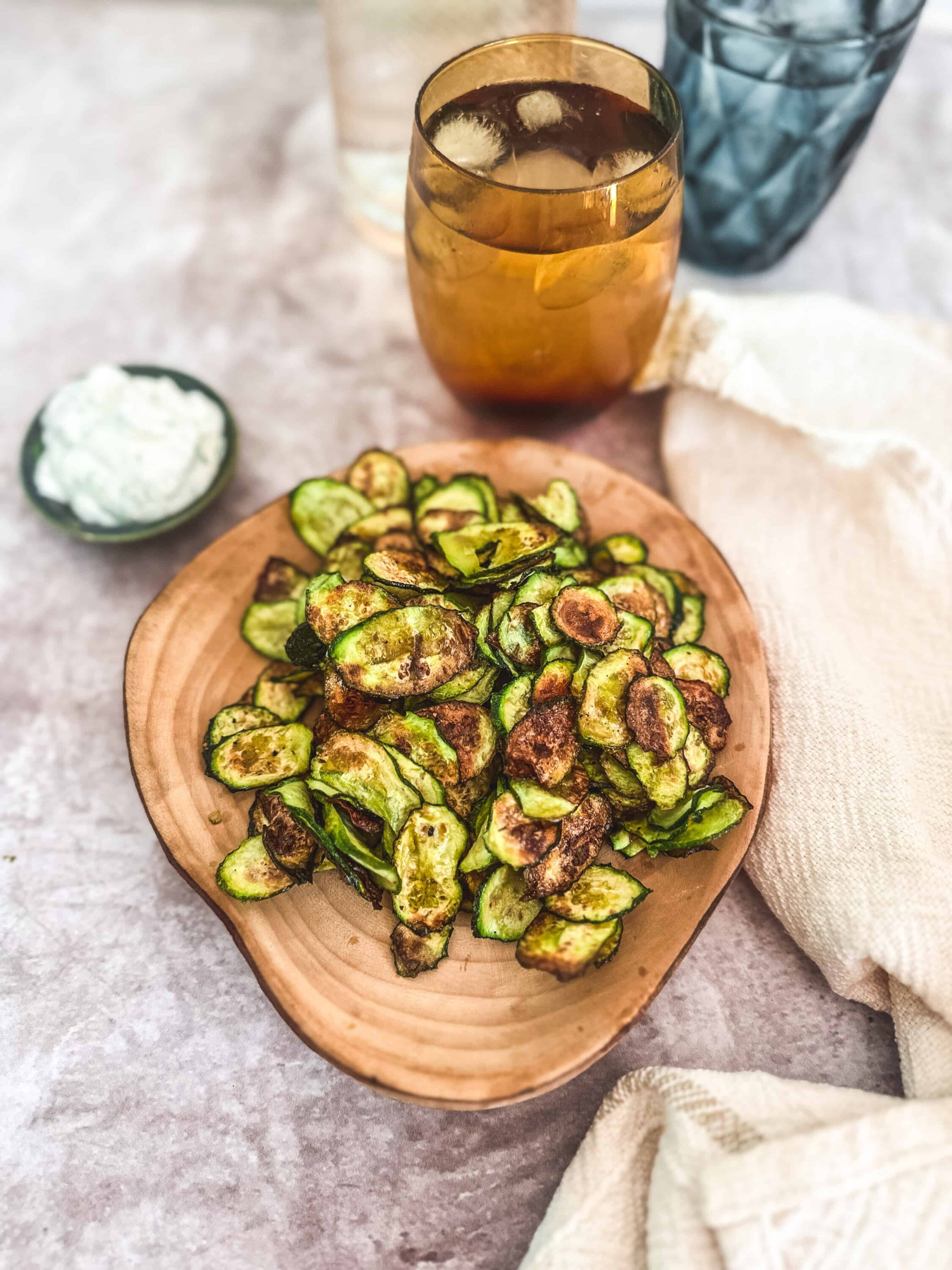 Crispy roasted zucchini chips served on a wooden platter with a side of creamy dip. Refreshing iced tea in a gold glass with water and glasses nearby.