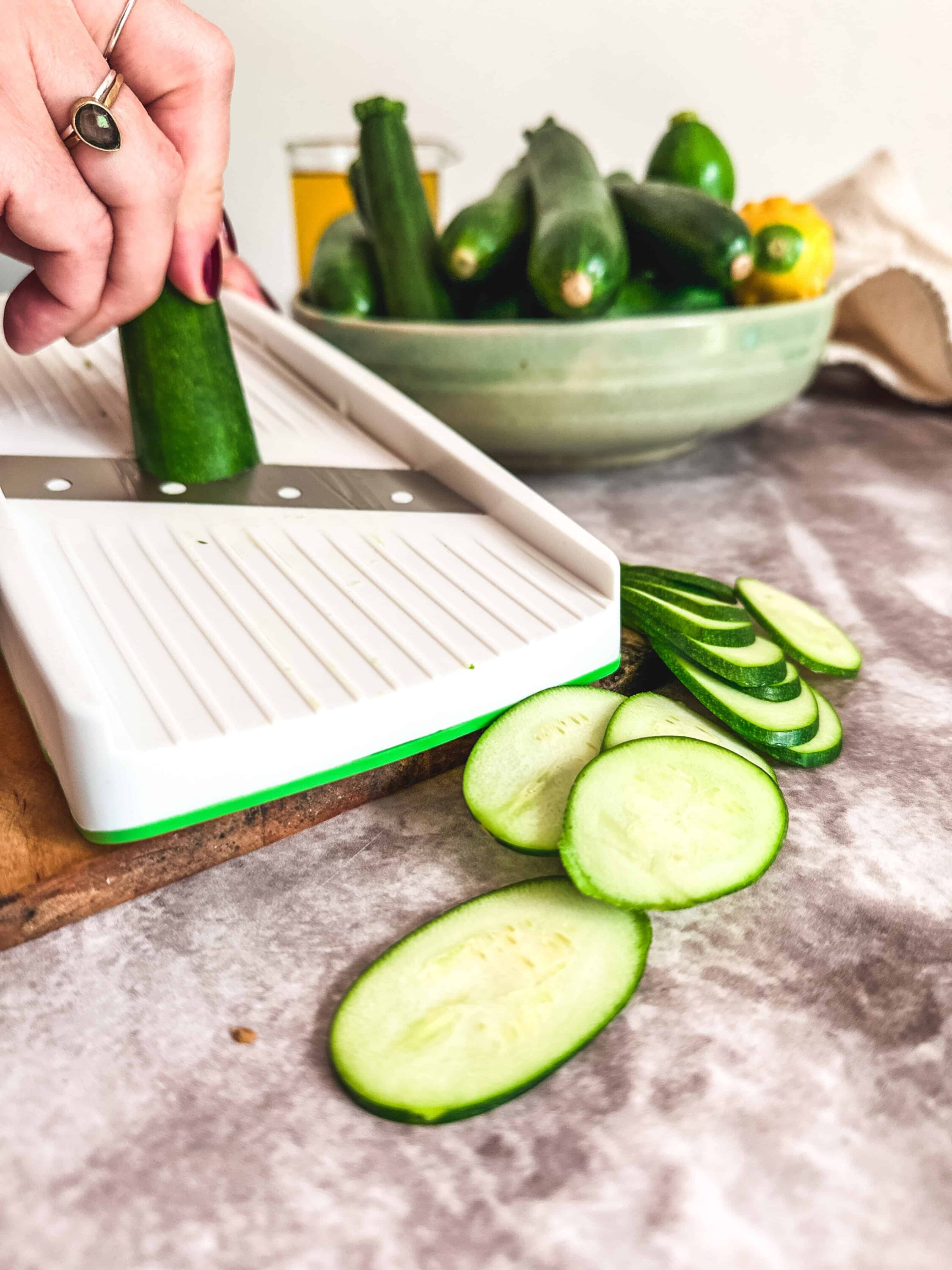 Slicing fresh cucumbers on a cutting board with a mandoline, with a bowl of zucchinis and a yellow squash in the background.