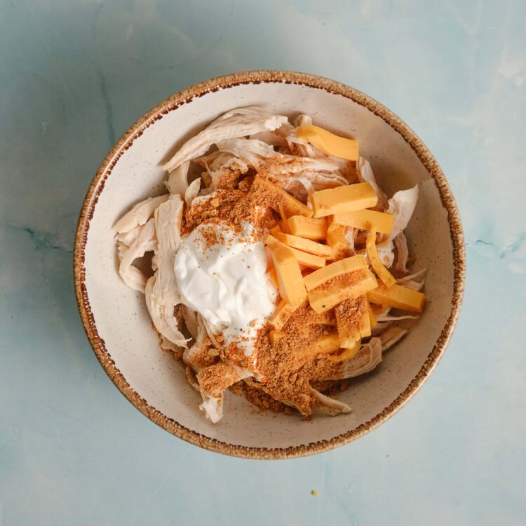 Shredded chicken with cheese, sour cream, and spices in a bowl, ready for cooking or serving.