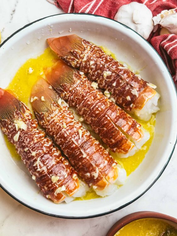 Succulent lobster tails drizzled with buttery garlic sauce, garnished with minced garlic and served in a white dish.
