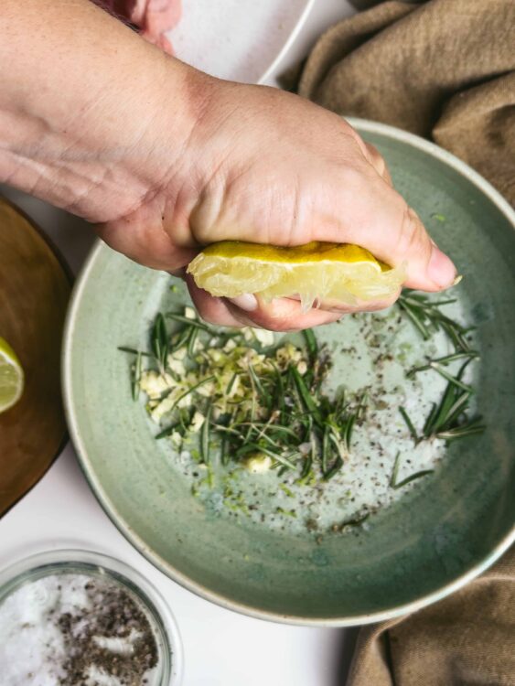 Fresh lemon being squeezed over chopped garlic and rosemary in a green bowl for flavorful cooking.