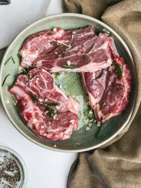 Seasoned raw lamb chops with garlic and rosemary in a ceramic dish for cooking or marinating.