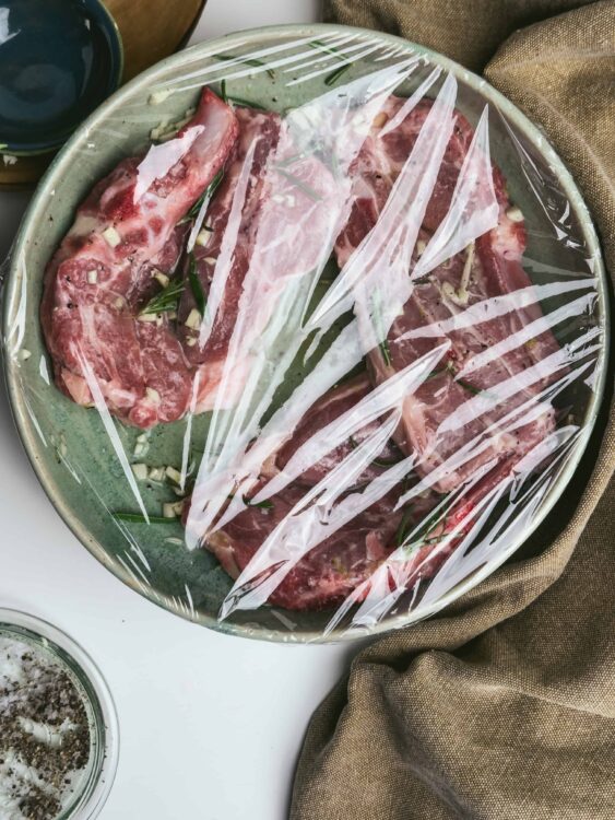 Marinated raw pork ribs with garlic and herbs, covered with plastic wrap in a baking dish.