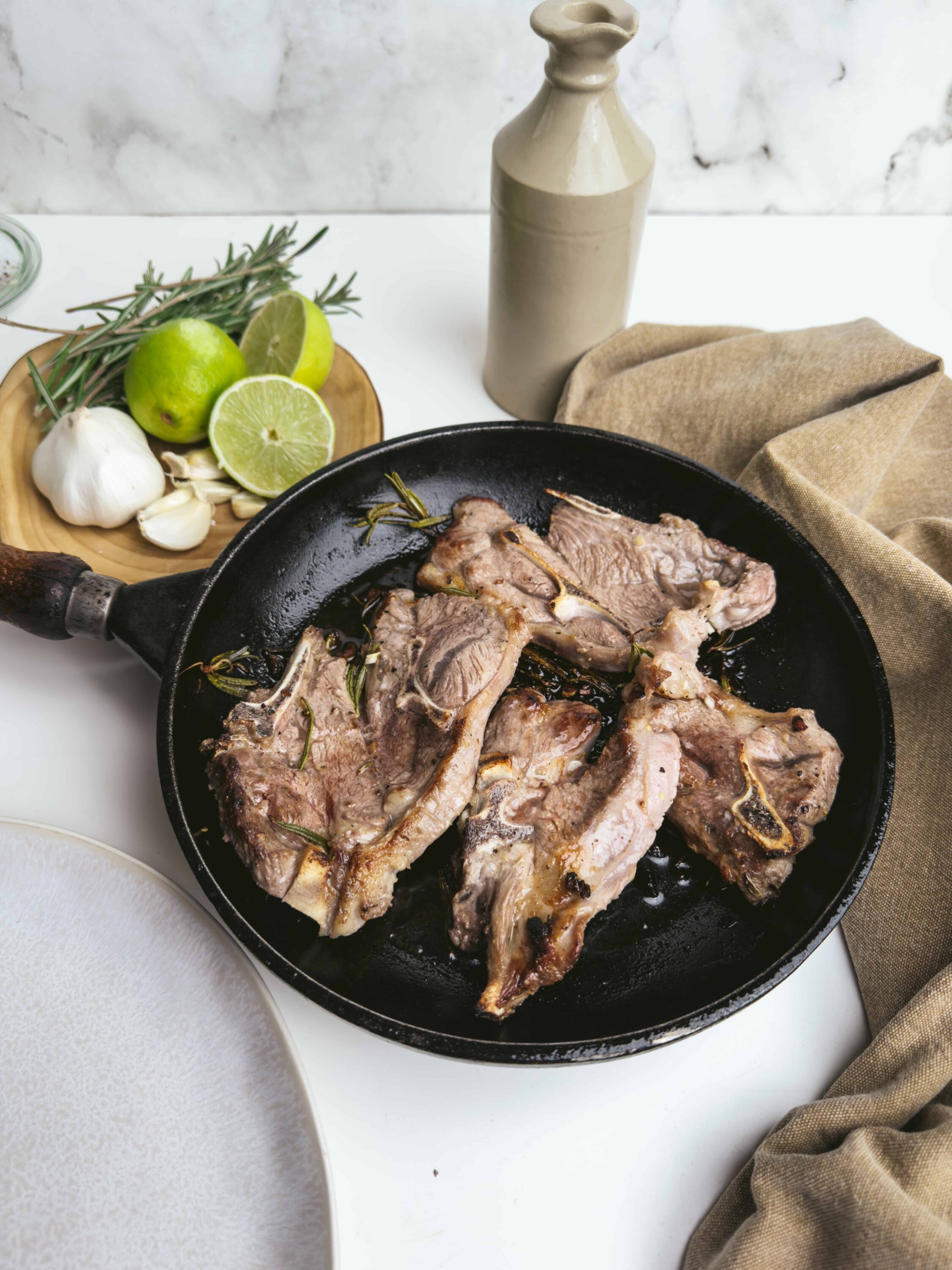 Juicy grilled lamb chops in a cast iron skillet with herbs, garlic, and olive oil on a white kitchen countertop.