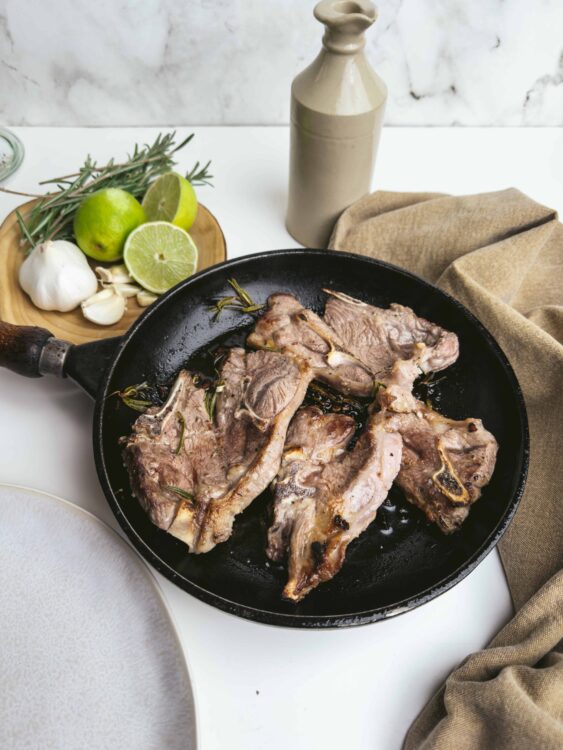 Juicy grilled lamb chops in a cast iron skillet with herbs, garlic, and olive oil on a white kitchen countertop.