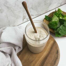 Creamy ranch dressing with a wooden stir stick on a round wooden serving board. Fresh lettuce in a wooden bowl next to the dressing.
