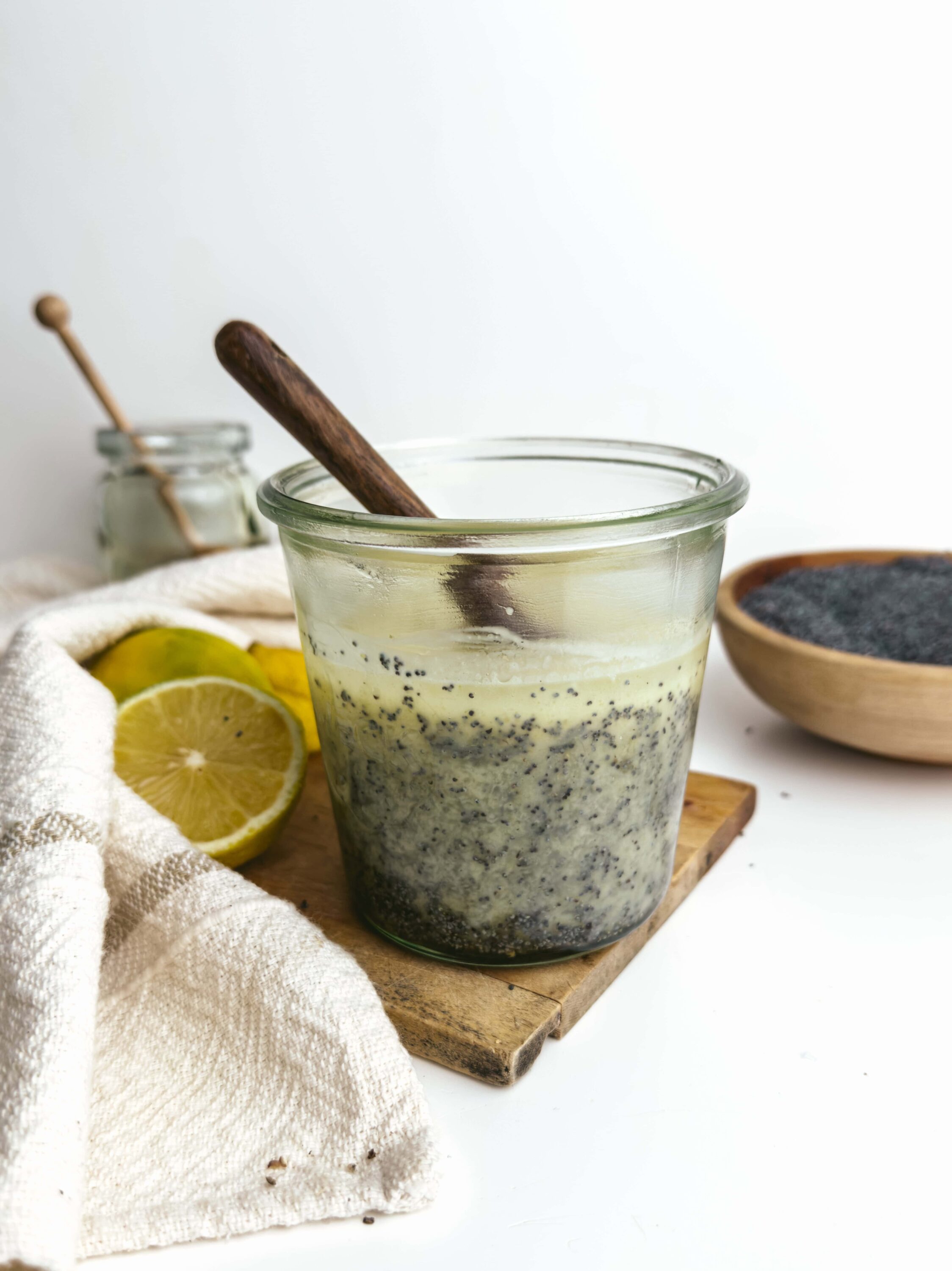 Lemon chia seed pudding with dark poppy seeds in glass jar, wooden spoon, lemon slices, and black sesame seeds on white background.