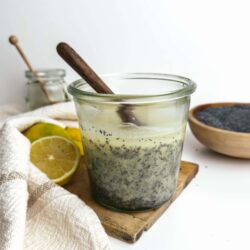 Lemon chia seed pudding with dark poppy seeds in glass jar, wooden spoon, lemon slices, and black sesame seeds on white background.