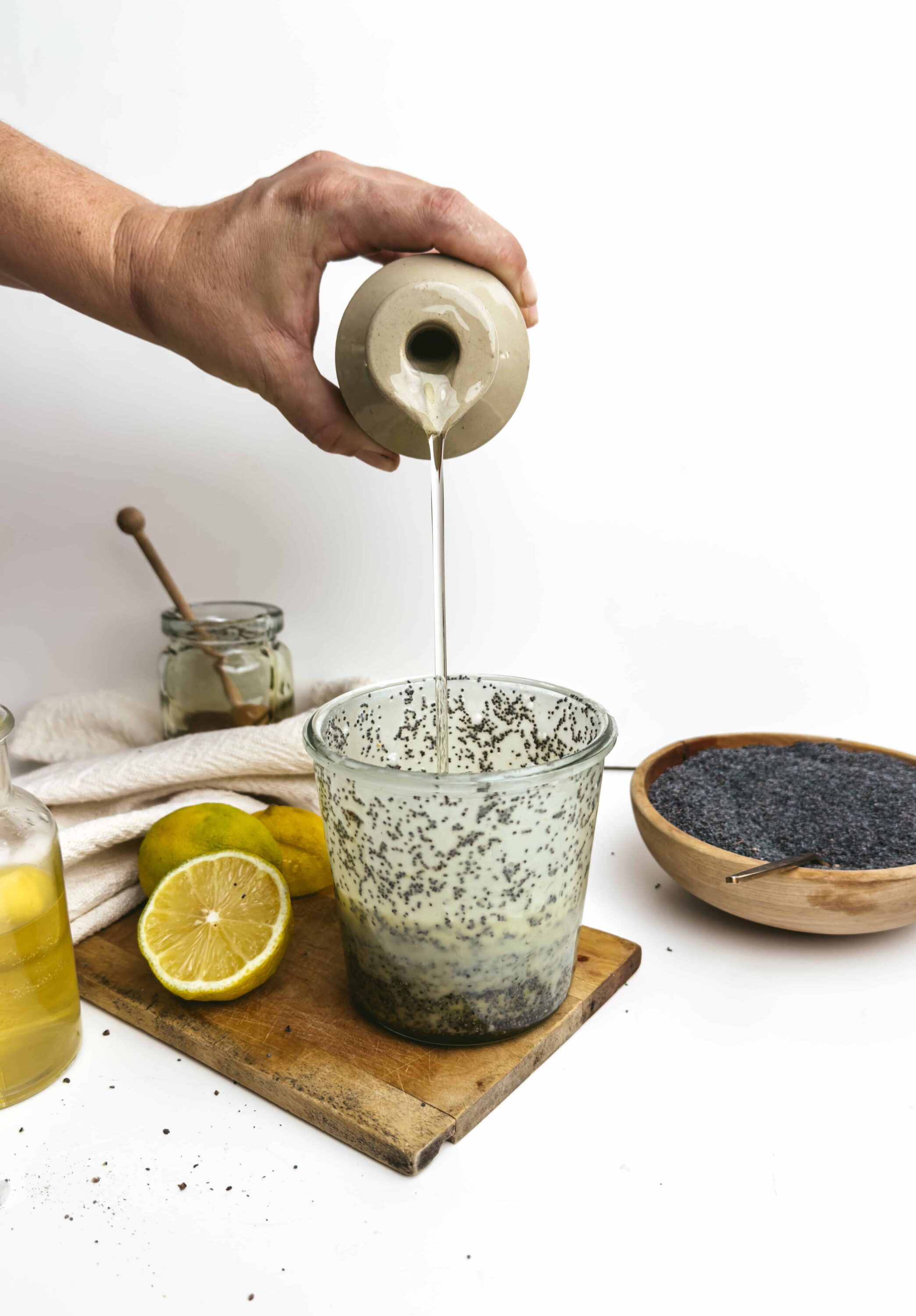 Smooth lemon poppy seed muffin batter being poured into a glass mixing bowl, with lemon slices and poppy seeds on a white background.