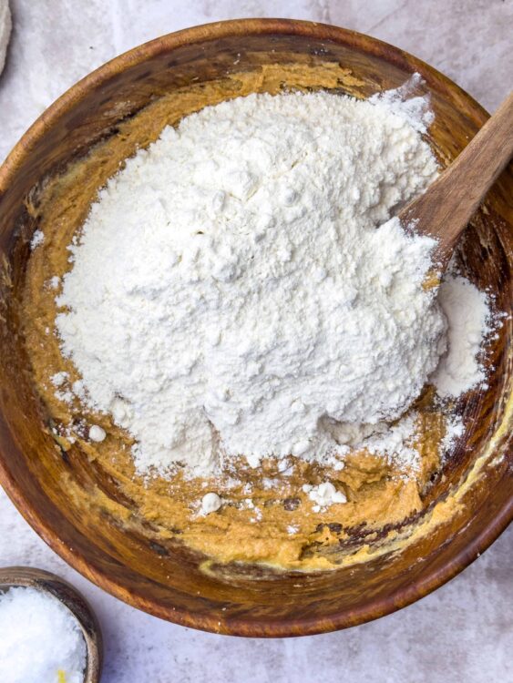 Flour being added to cookie dough in a rustic wooden mixing bowl for baking.