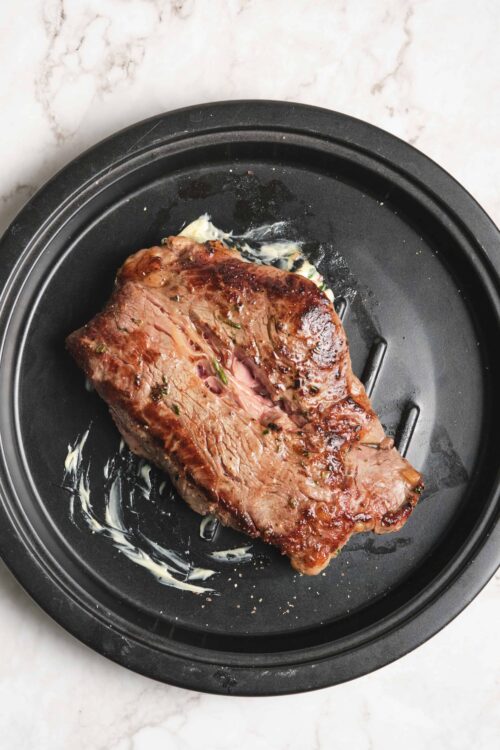Juicy, cooked steak on a black air fryer tray with melted butter, seasoned with herbs and spices, ready to serve.
