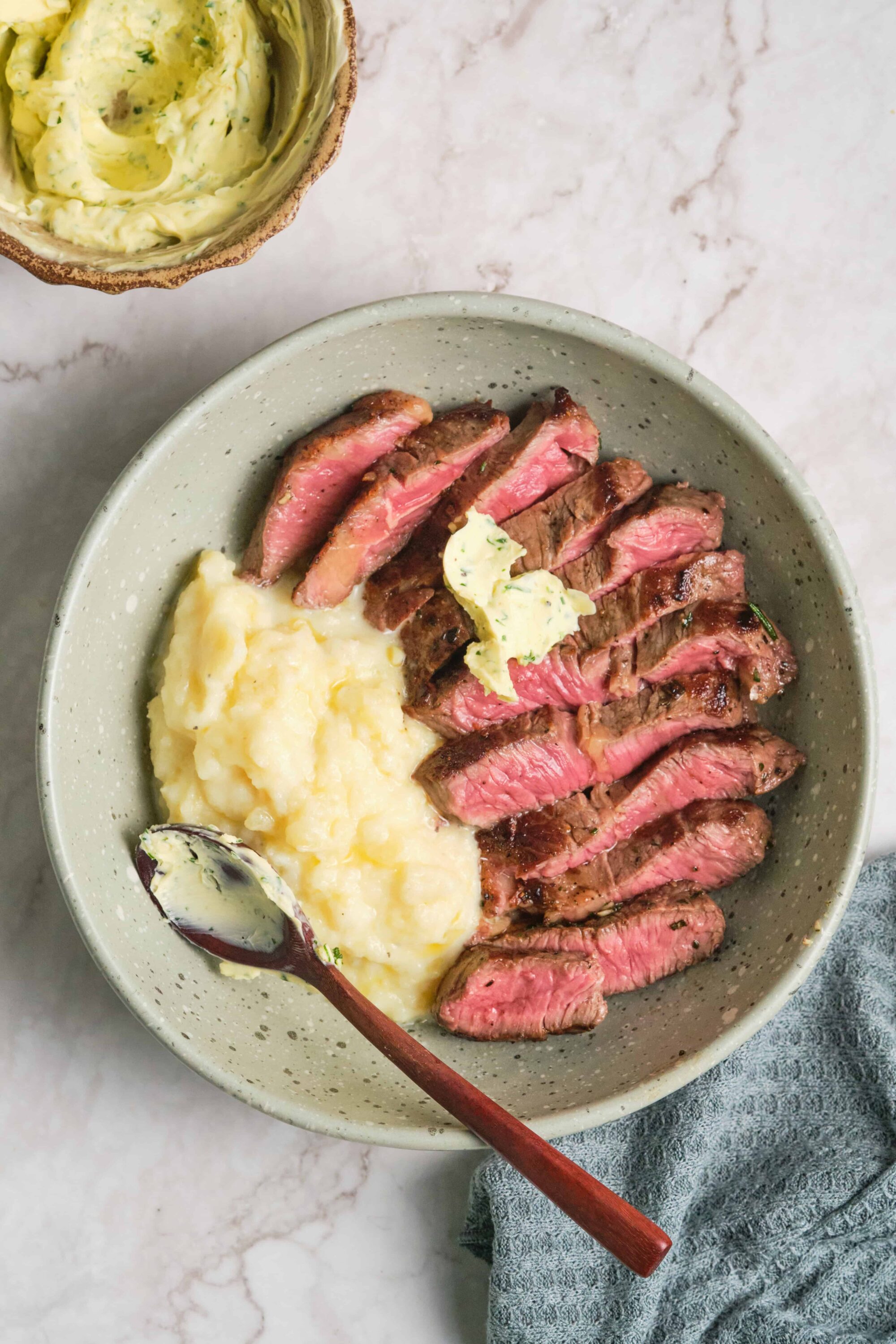 Juicy sliced steak with mashed potatoes and herb butter, served in a ceramic bowl on a marble surface.