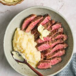 Juicy sliced steak with mashed potatoes and herb butter, served in a ceramic bowl on a marble surface.