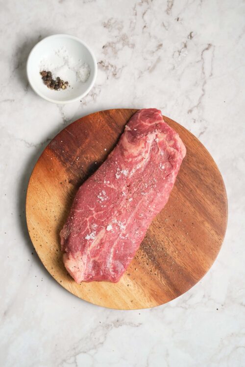Salt and pepper seasoned raw beef steak on wooden cutting board over white marble surface.