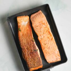 Perfectly cooked salmon fillets searing in a black skillet on a light marble countertop.