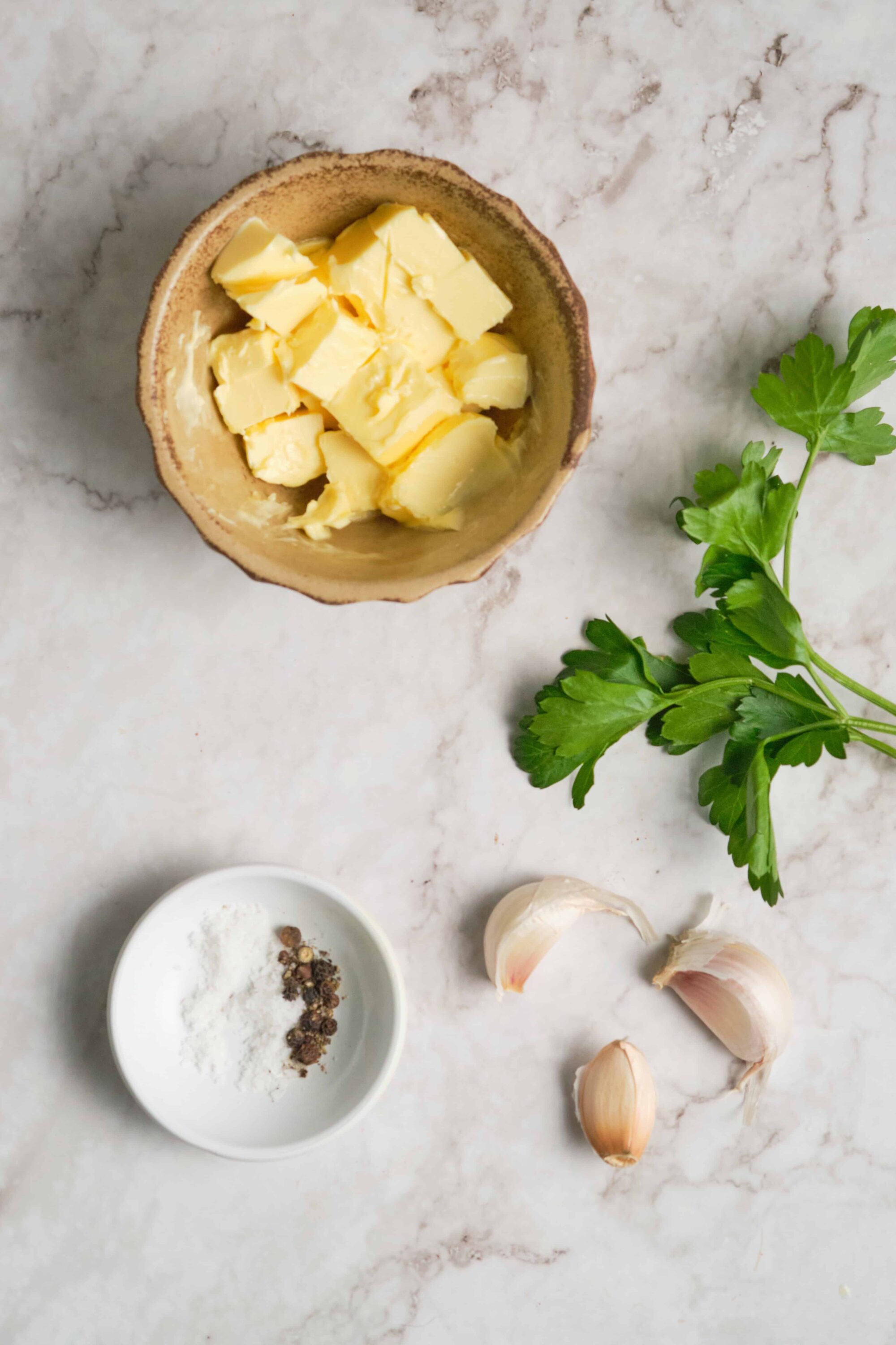 Creamy butter cubes in a rustic dish, garlic cloves, parsley, and seasoning on marble surface for delicious baking.