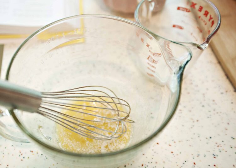 Baked Bree, mixing ingredients for baking, whisk in glass bowl with measuring jug on speckled countertop.