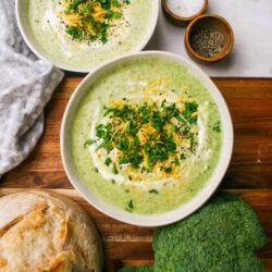 Creamy broccoli cheese soup served in bowls garnished with herbs and shredded cheese. Fresh bread and broccoli on a rustic wooden table.