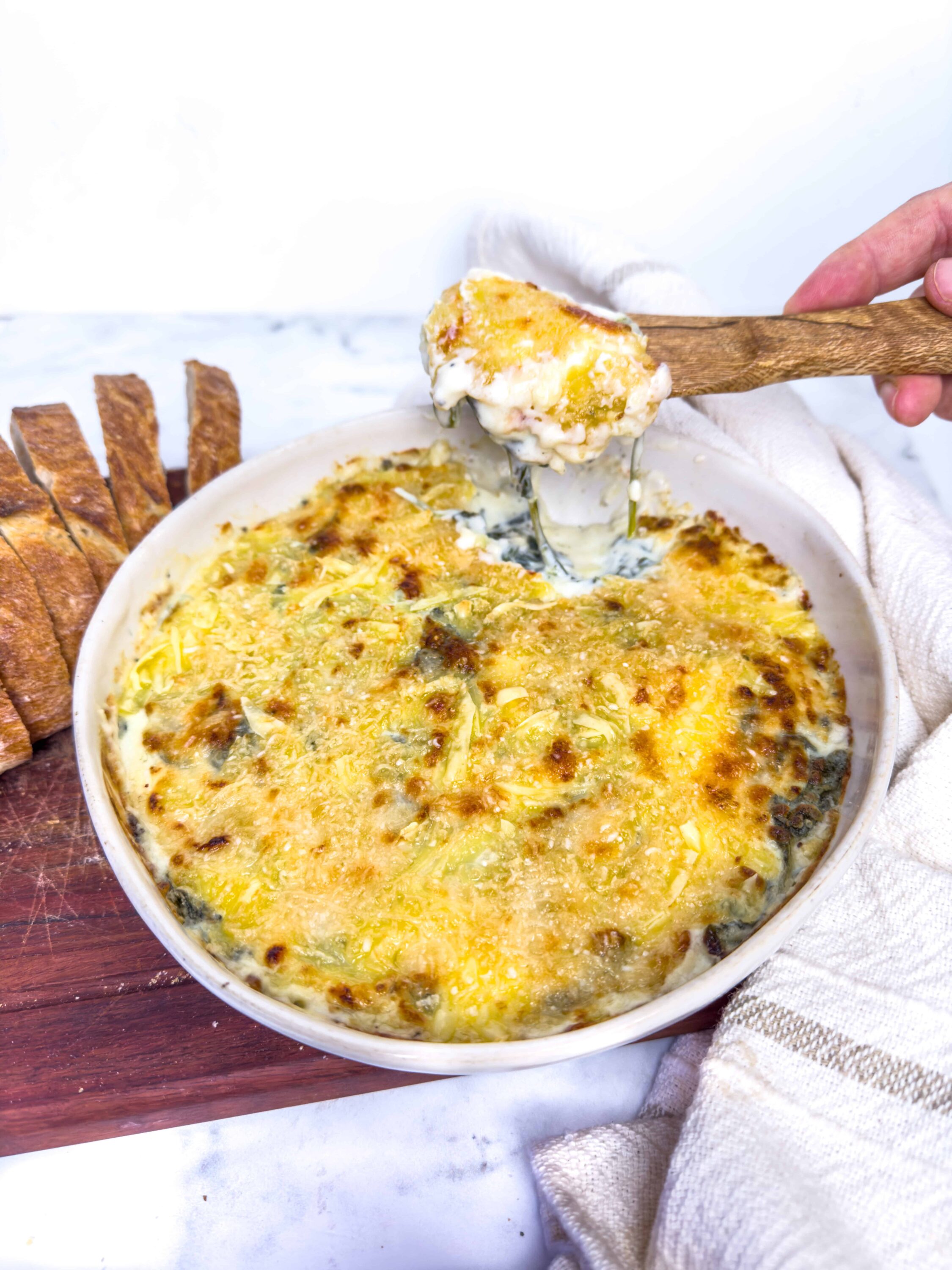 Creamy baked cheesy dip with vegetables in a white ceramic dish, with toasted bread slices on the side.