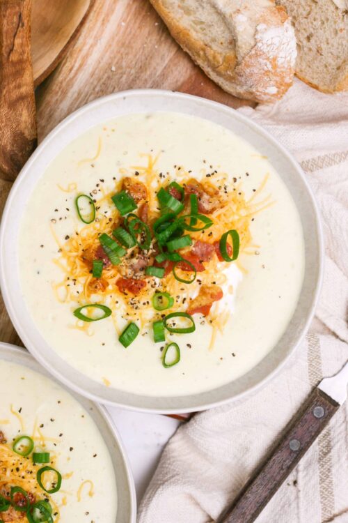 Creamy potato soup garnished with bacon, shredded cheddar, and green onions on a rustic table.