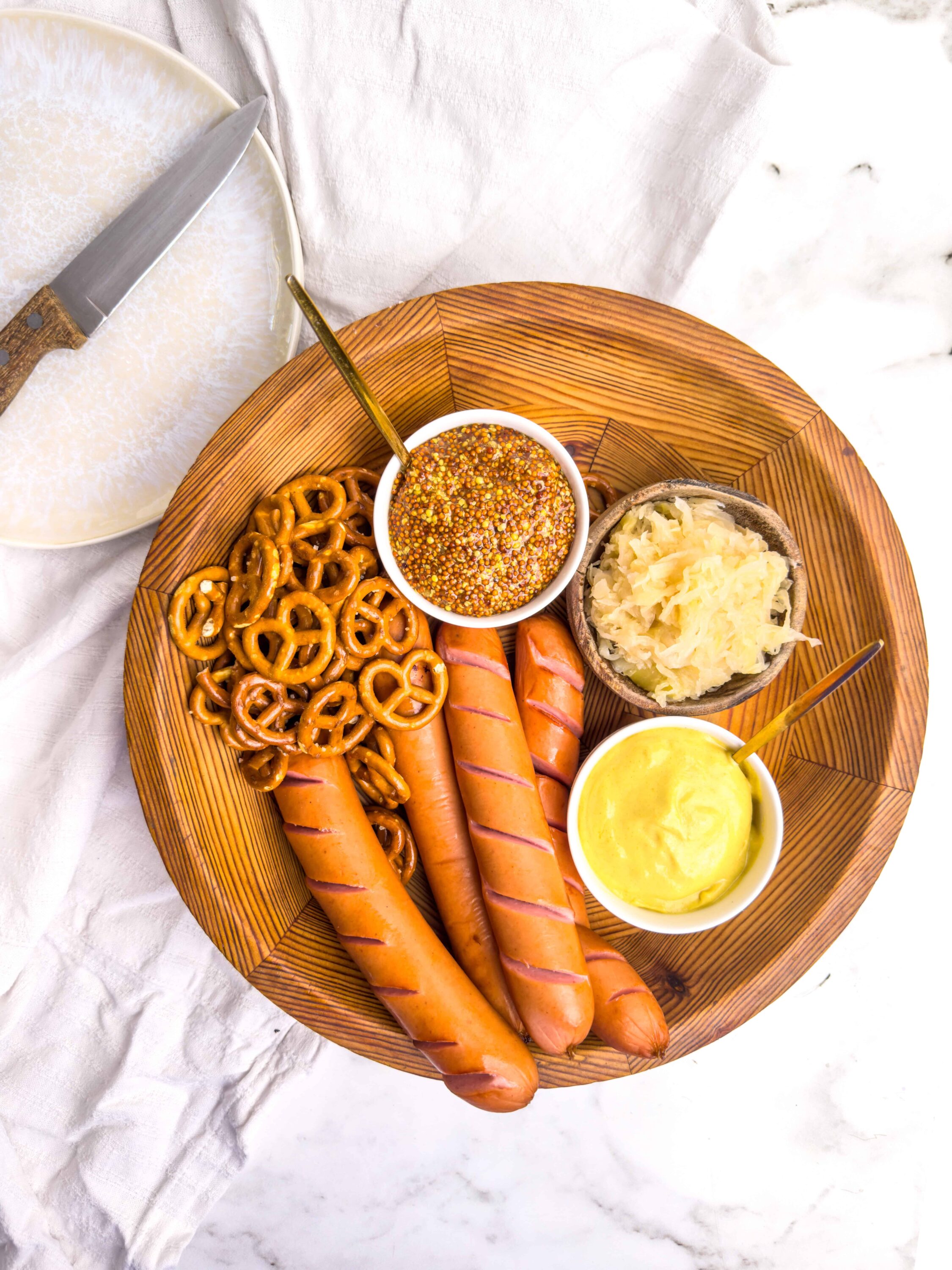 Grilled hot dogs with pretzels, mustard, sauerkraut, and potato salad on a wooden platter, perfect for summer cookouts.