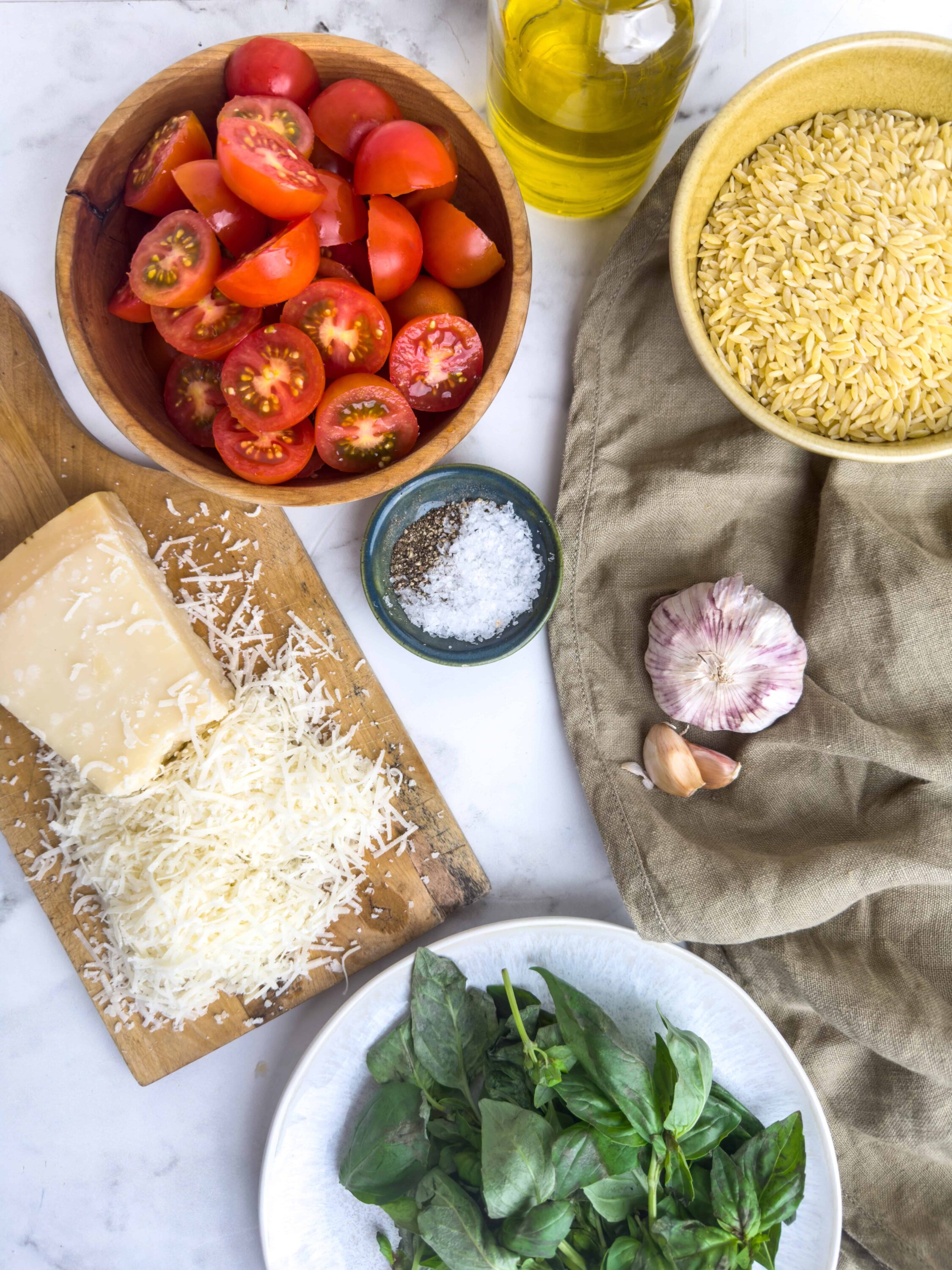 Cherry tomatoes on a wooden bowl, fresh basil leaves, grated cheese, garlic, olive oil, orzo pasta, and seasonings arranged on a marble surface for pasta salad preparation.