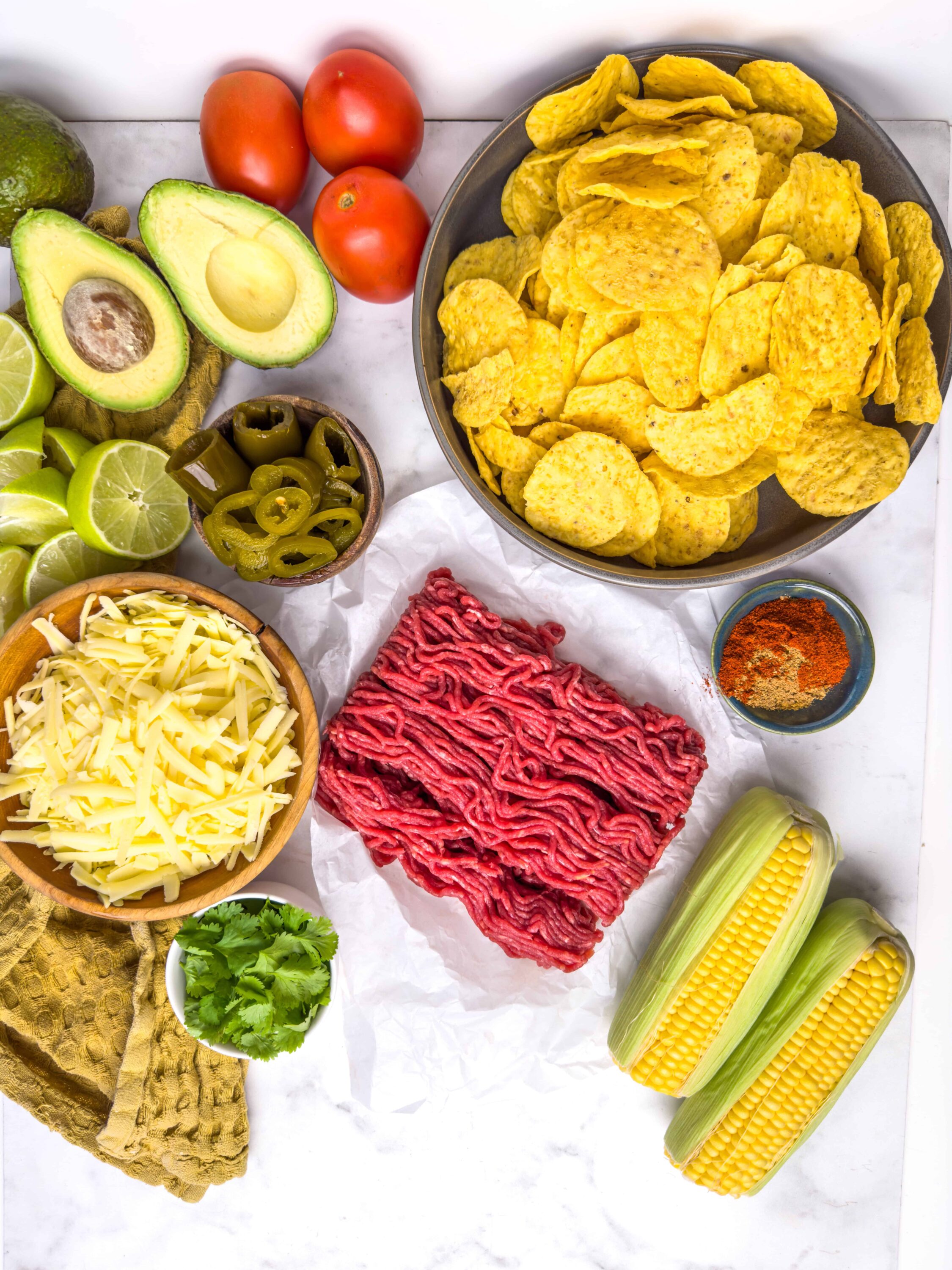 Fresh taco ingredients including ground beef, shredded cheese, corn, avocado, tomatoes, jalapenos, lime, corn tortillas, and potato chips.