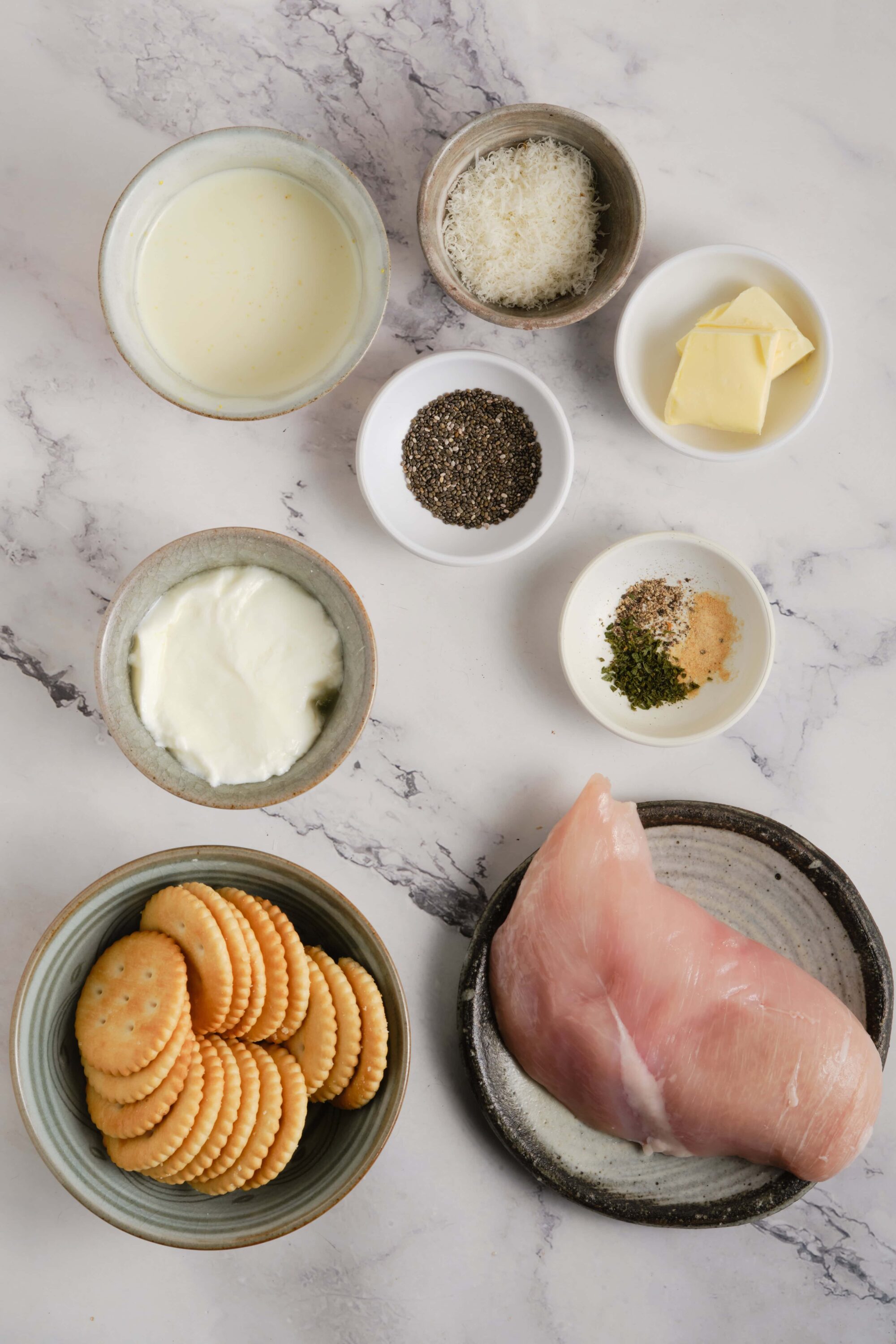 Cream cheese and chicken ingredients for a baked dish, with saltine crackers and seasoning, on a marble surface.