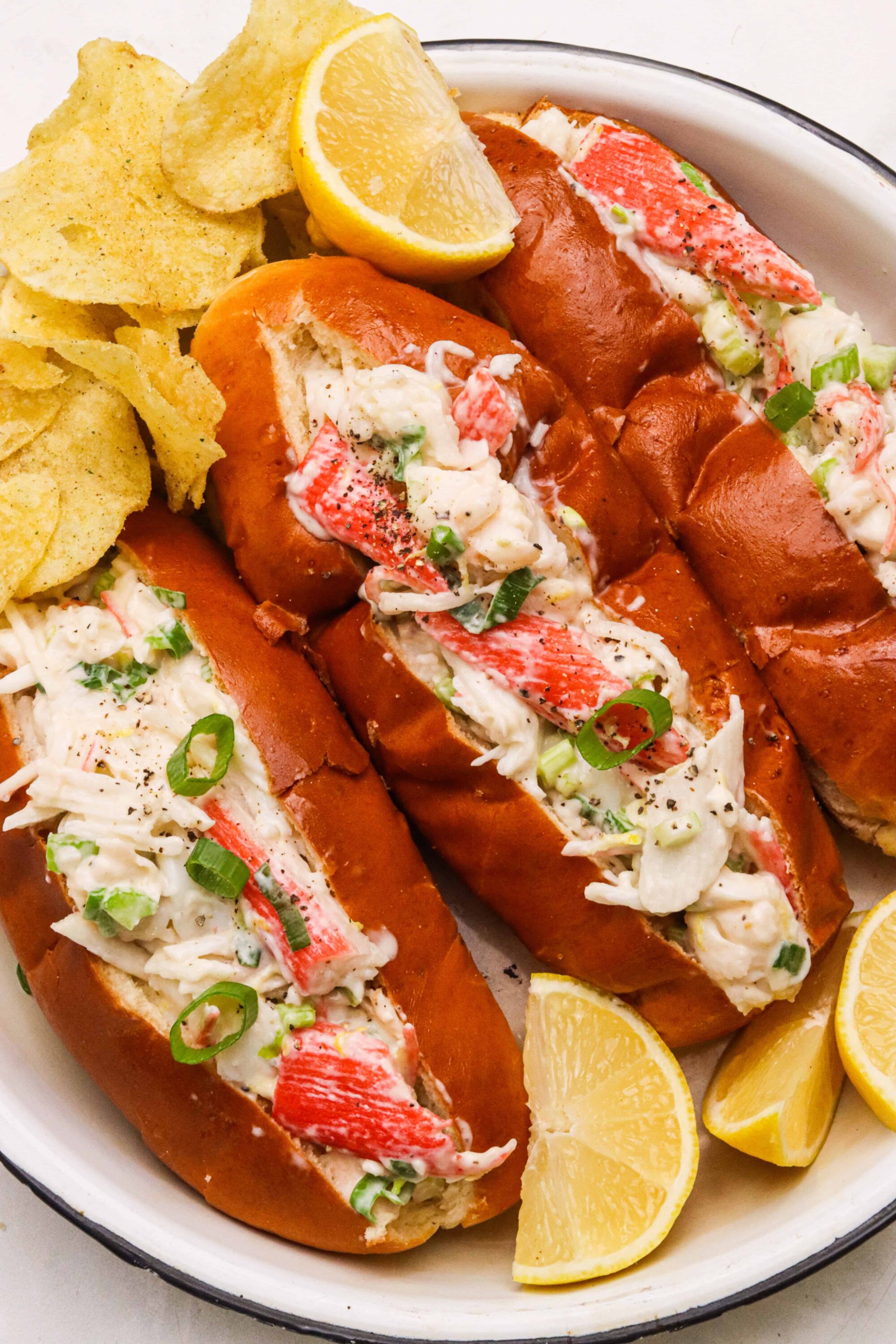 Juicy crab salad rolls with fresh vegetables in toasted hot dog buns. Served with potato chips and lemon wedges for a delicious seafood meal.