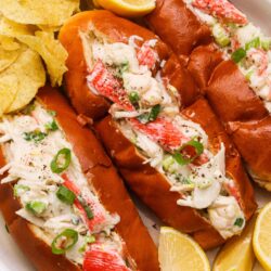 Juicy crab salad rolls with fresh vegetables in toasted hot dog buns. Served with potato chips and lemon wedges for a delicious seafood meal.
