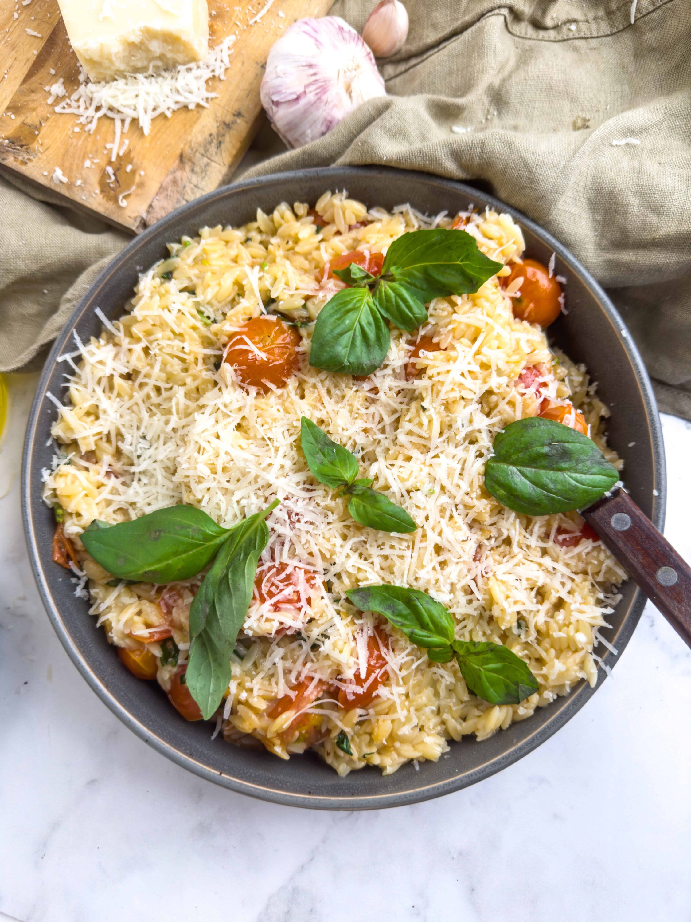 Creamy orzo pasta with cherry tomatoes, fresh basil, and grated cheese in a gray skillet.