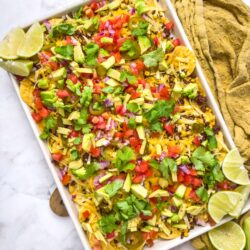 Colorful loaded nachos topped with fresh avocado, jalapenos, tomatoes, onions, and cilantro on a white platter.