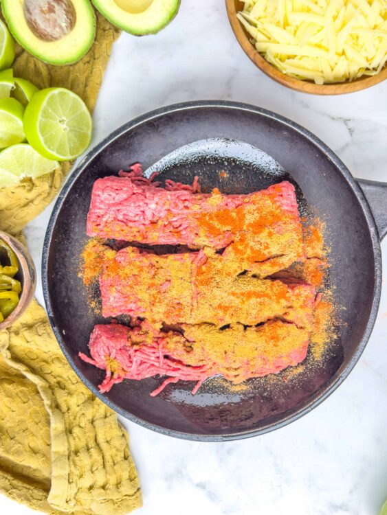 Raw ground beef in a skillet with seasoned spices, surrounded by fresh lime and avocado, ready for cooking.