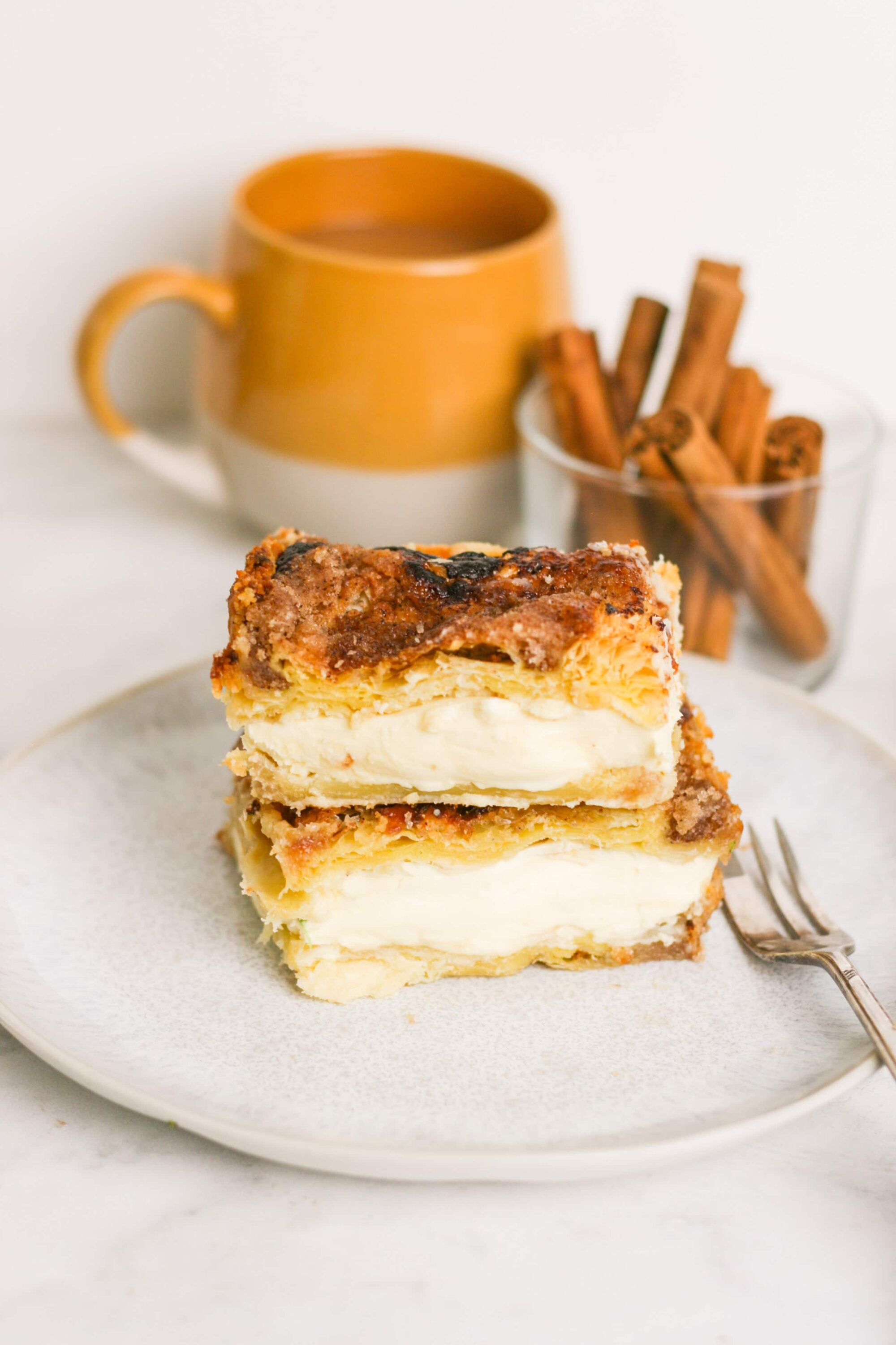 Cream cheese carrot cake slice on white plate with cinnamon sticks and coffee mug in background.