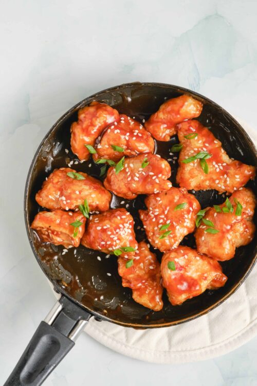 Sweet and spicy chicken bites with sesame seeds and green onions on a black skillet, served as a delicious appetizer or meal.