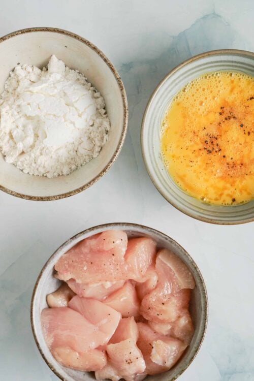 Flour, beaten eggs, and raw chicken on a white surface, ingredients for baked chicken recipe.
