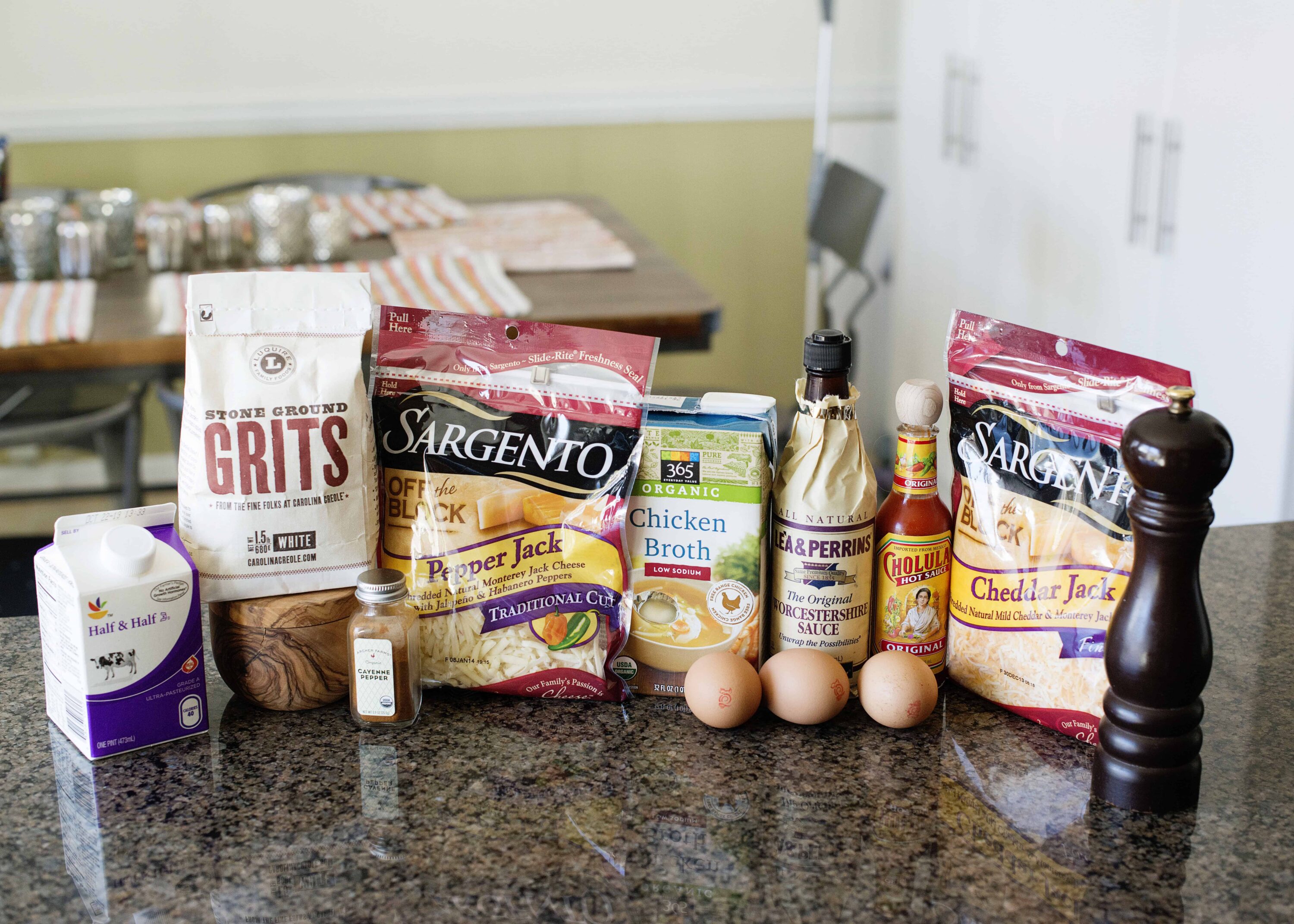 Fresh baking ingredients including grits, cheese, chicken broth, eggs, and hot sauce on a kitchen countertop.