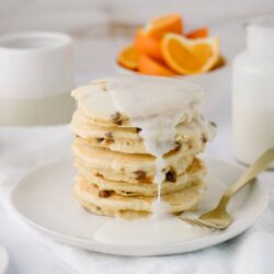 Fluffy chocolate chip pancakes with creamy white glaze, served on a white plate with fresh orange slices in the background.