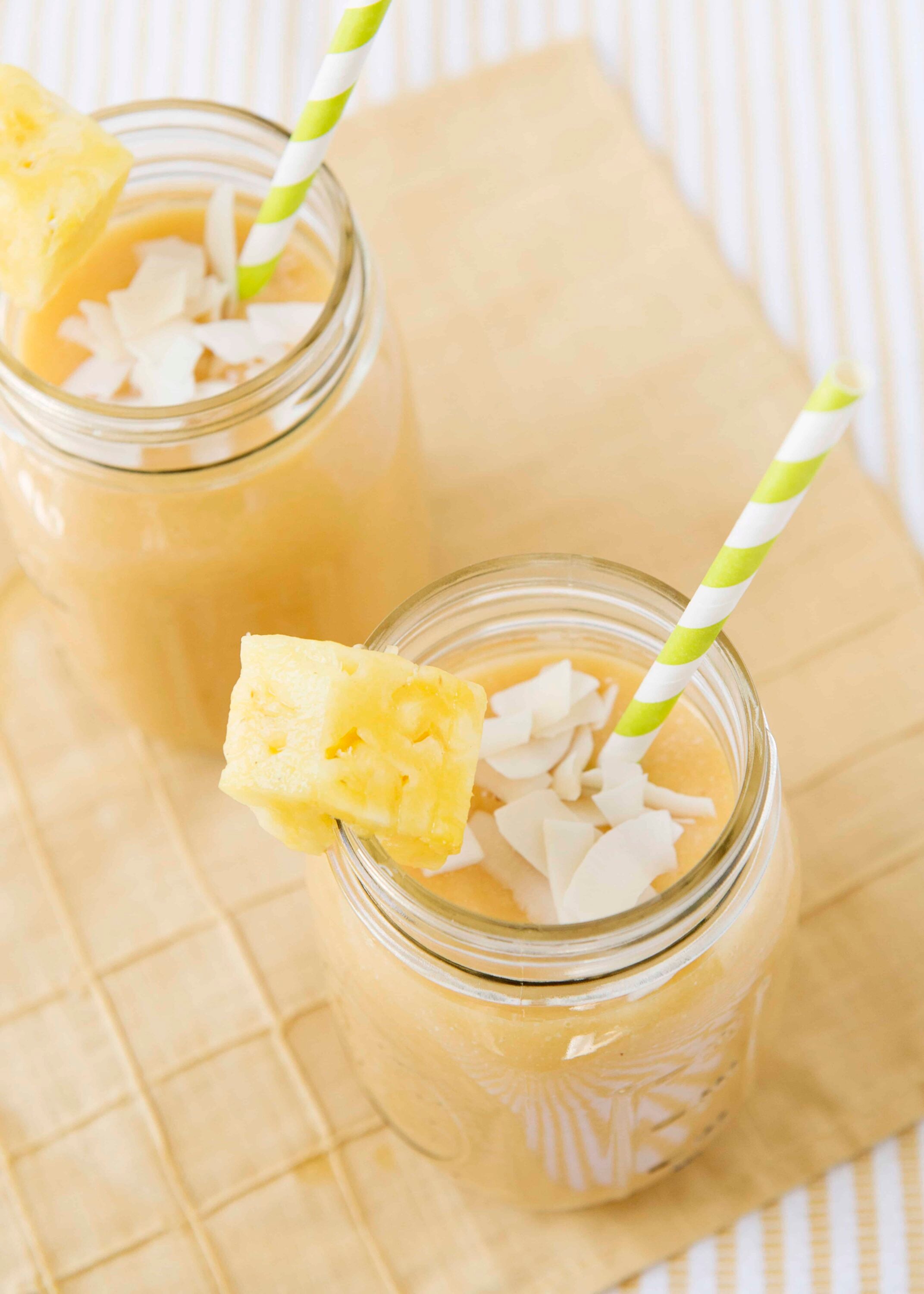Pineapple and coconut smoothies in glass jars with striped straws, garnished with pineapple chunks and coconut flakes.