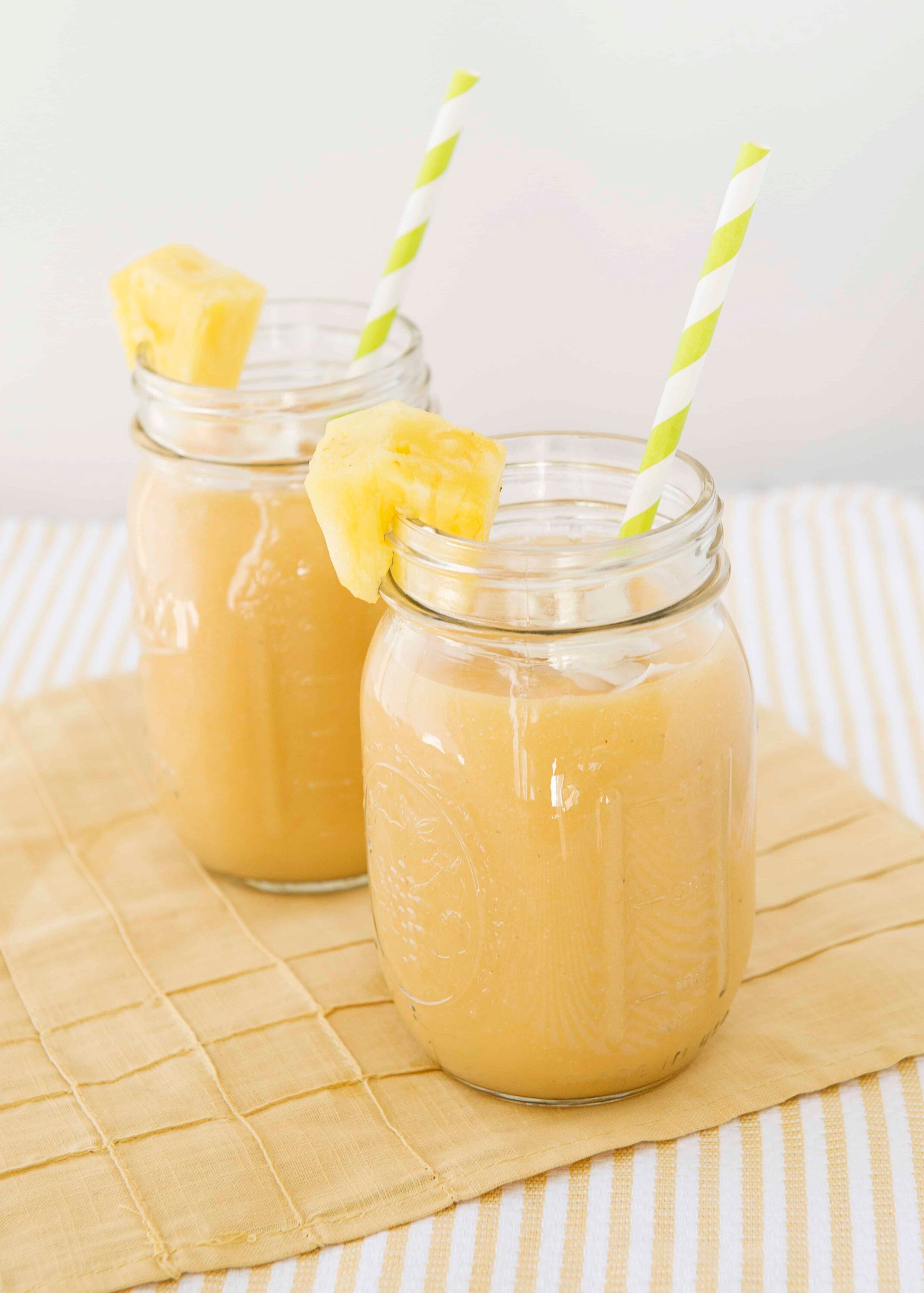Creamy pineapple smoothie served in mason jars with striped straws and pineapple garnish. Refreshing summer beverage perfect for breakfast or snack.