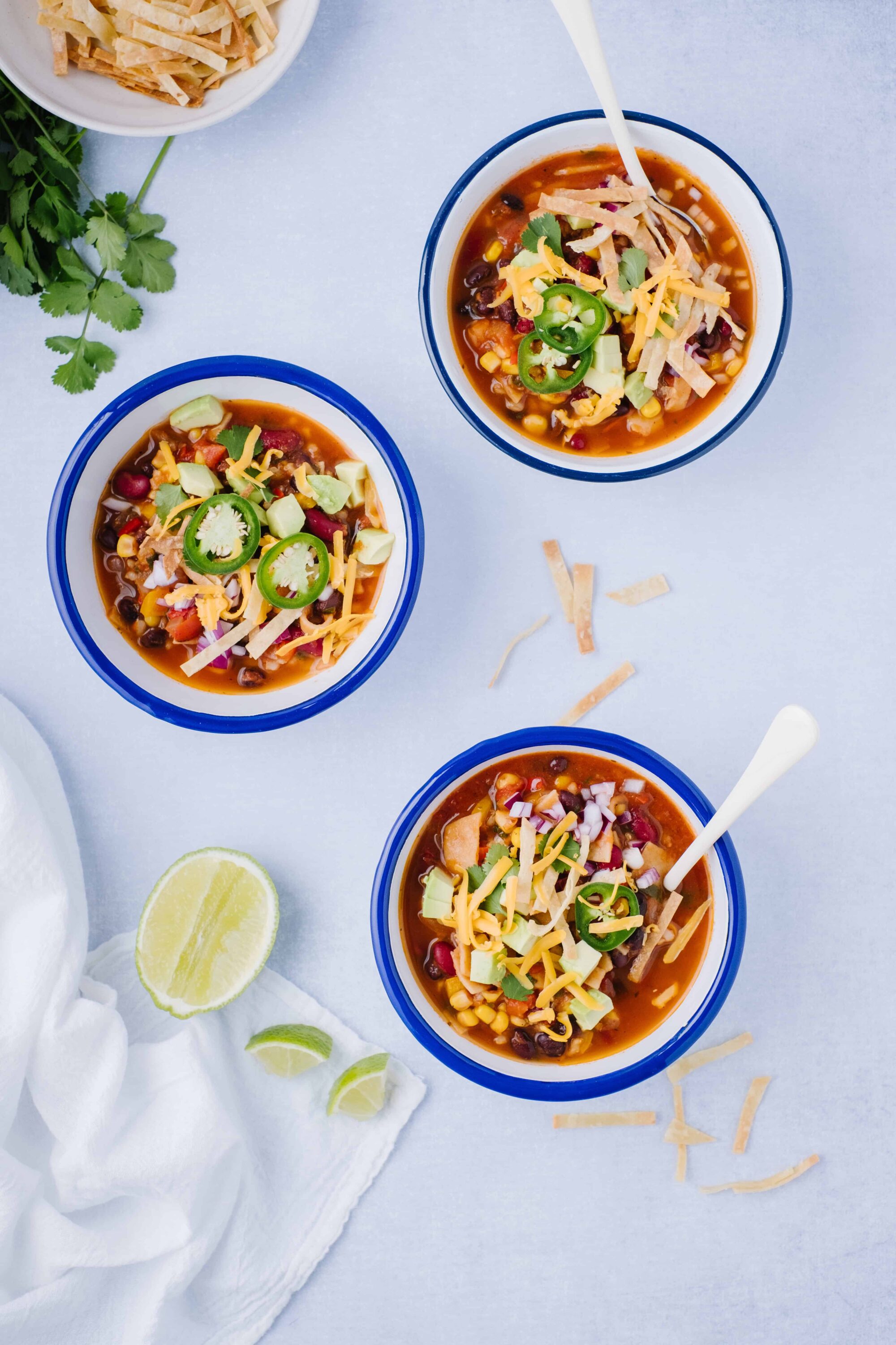Cheesy beef chili served in blue bowls with fresh toppings and lime wedges on a white background.
