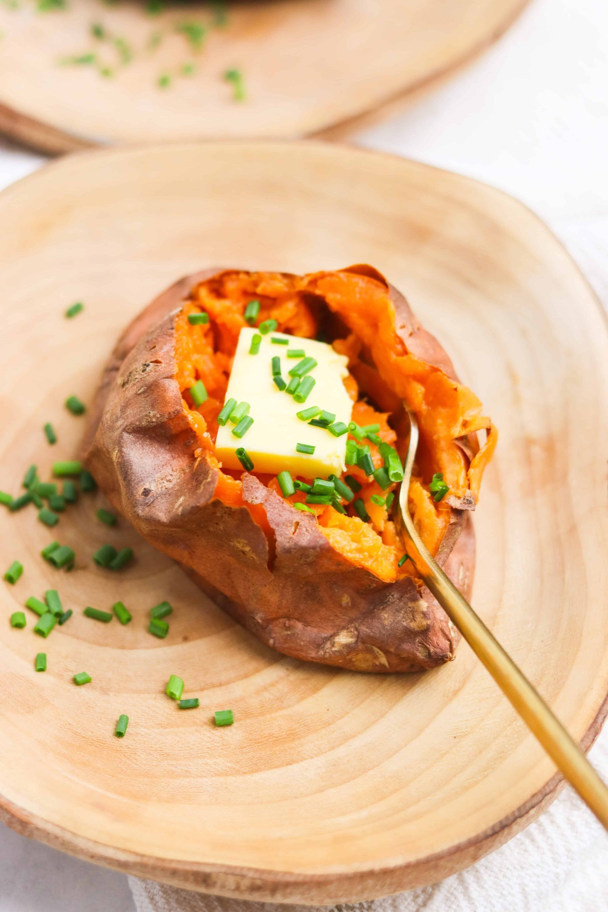 Baked sweet potato topped with butter and chopped chives on a wooden plate.