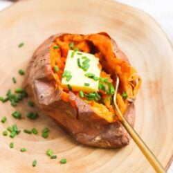 Baked sweet potato topped with butter and chopped chives on a wooden plate.