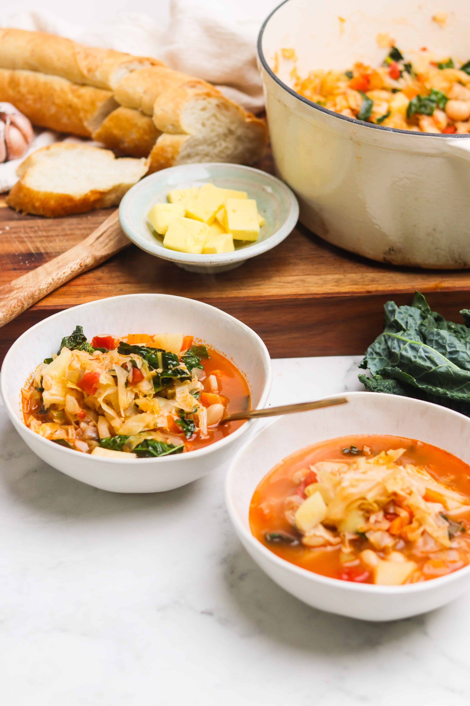 Savory vegetable soup with shredded cabbage in white bowls, fresh bread, and butter on a wooden board.
