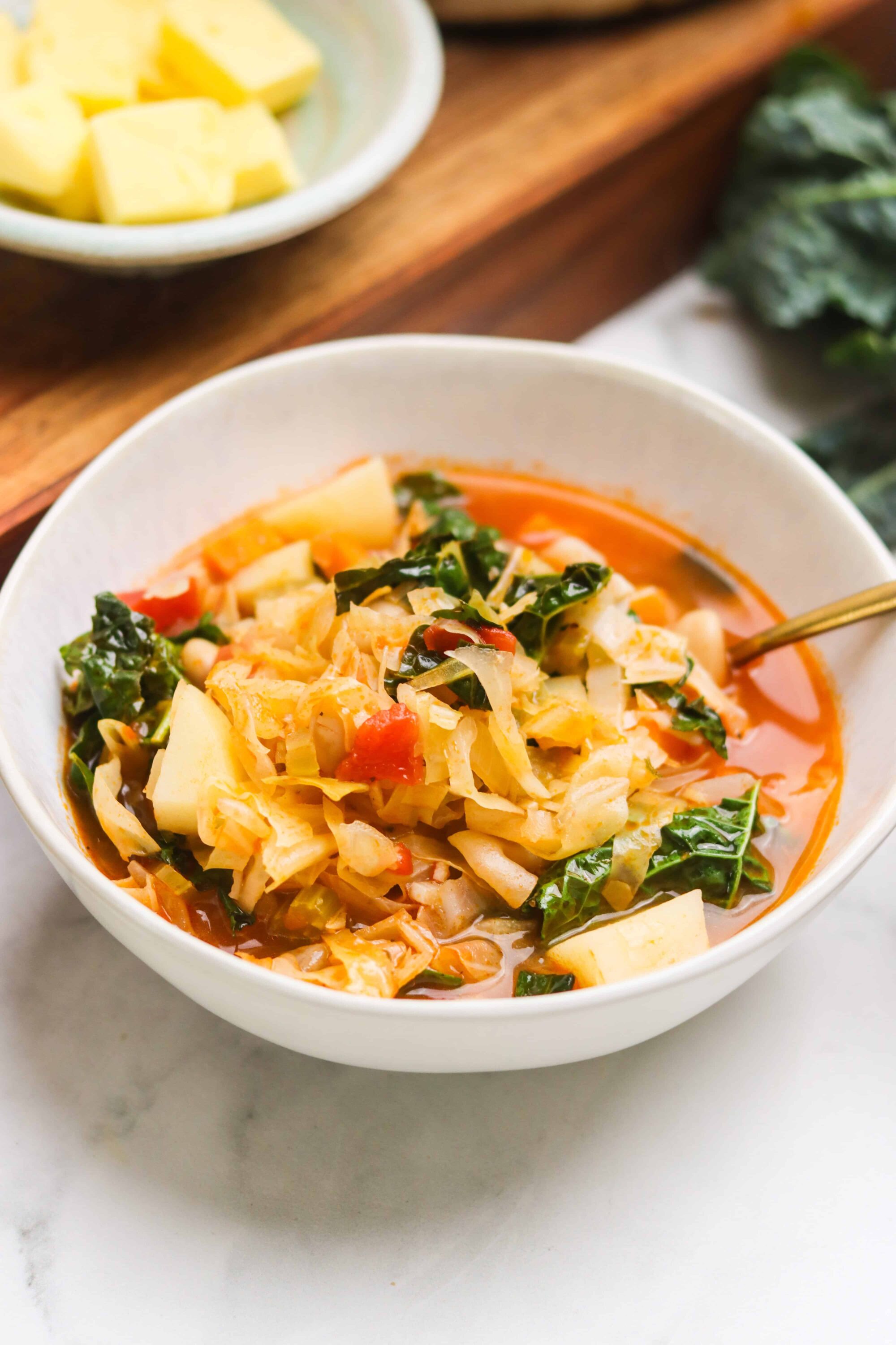 Savory cabbage soup with vegetables in a white bowl, fresh ingredients on baking tray, cozy home-cooked meal.